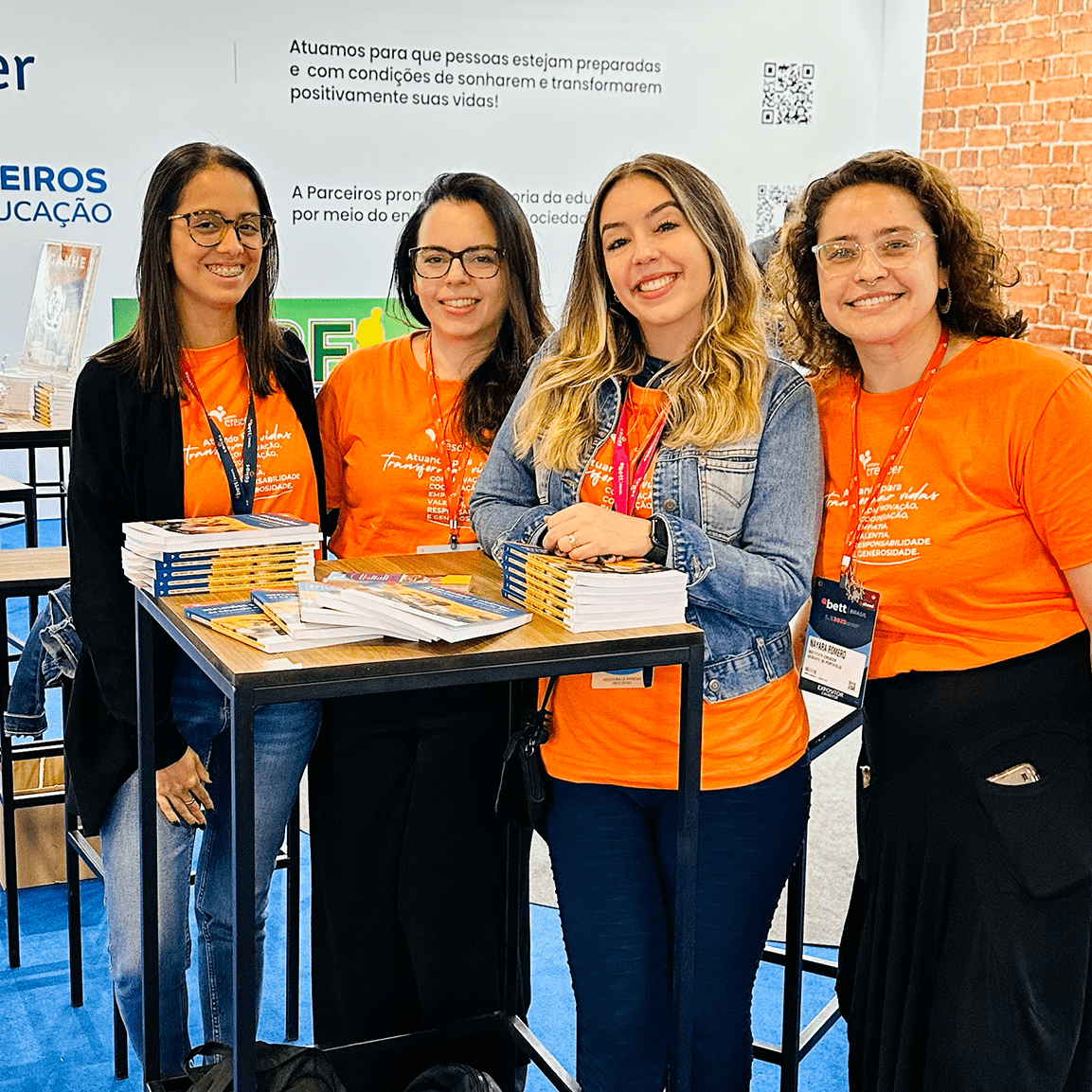 Grupo de quatro mulheres usando camisetas laranjas ao lado de uma mesa cheia de livros para ilustrar nosso sistema de atendimento "Fale conosco".