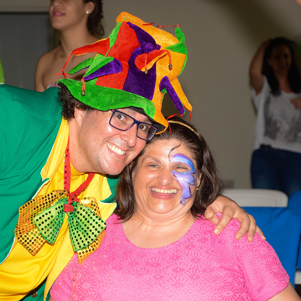 Foto de um homem vestido de palhaço abraçando uma mulher sorridente, usando uma camiseta rosa e com uma borboleta pintada em metade do rosto, em ação do projeto Conecta.