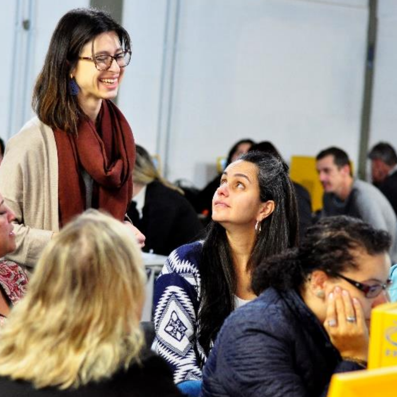 Uma mulher sorri calorosamente para um grupo de pessoas que estão sentadas em mesas realizando atividades do projeto Educonexão.